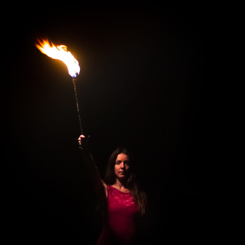 Olivia Fern wearing a red dress holds a burning torch in the dark night.